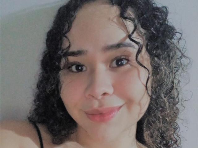 EveNailah tiene el pelo castaño rizado, ojos oscuros y una sonrisa sutil. Lleva puesto un top negro con tirantes finos, y apenas se ven sus pechos. Es una mujer atractiva de piel clara..