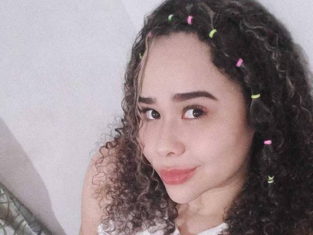 EveNailah tem cabelos castanhos cacheados com presilhas de cabelo rosa e verde, e está sorrindo com batom rosa, parecendo fofa e um pouco engraçada. Seus seios não estão visíveis..