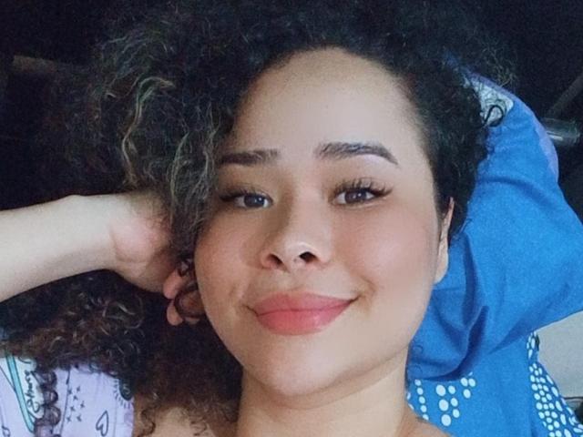 EveNailah a les cheveux bruns bouclés, une peau brun clair et un petit nez, et elle sourit avec ses lèvres roses. Elle est allongée, portant une chemise bleue et avec un bras levé..