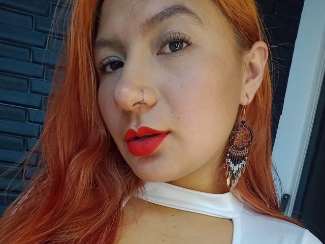 AradiaX with red lipstick, nose ring, and dangling earring, has orange hair and a white top, showing her face and upper chest, with a slight smile.