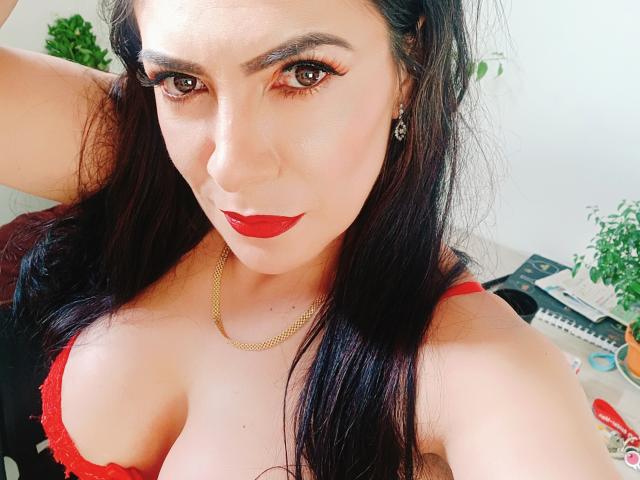 JoTaylor with long black hair, red lipstick, and red bra, showing her tits, wearing a gold chain, looking at the camera with eyeliner and a confident expression.
