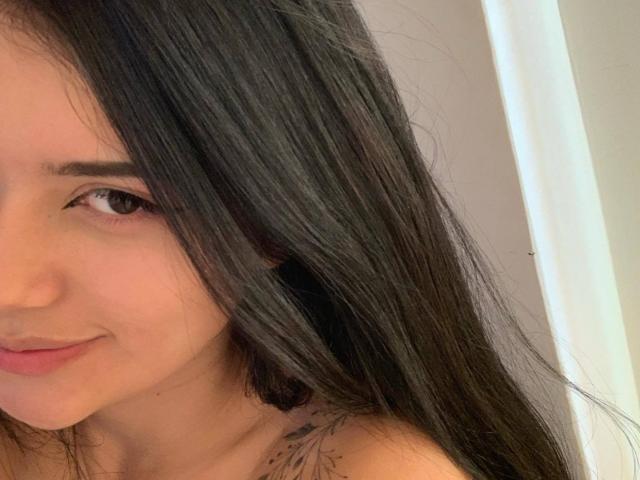 Het gezicht van HelenTay, met lang zwart haar, bruine ogen en een lichte glimlach. Ze heeft een kleine tatoeage op haar rechter schouder. Haar borsten zijn gedeeltelijk zichtbaar..