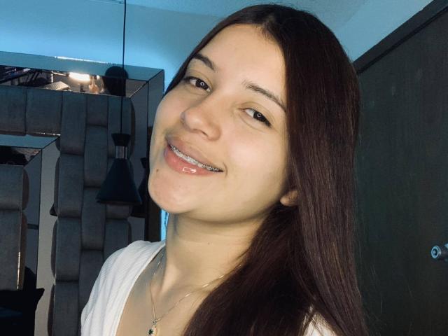 HelenTay, com cabelo castanho liso, sorrindo com a boca ligeiramente aberta, mostrando aparelhos ortodônticos de metal, vestindo uma blusa branca, em um quarto com pouca luz..