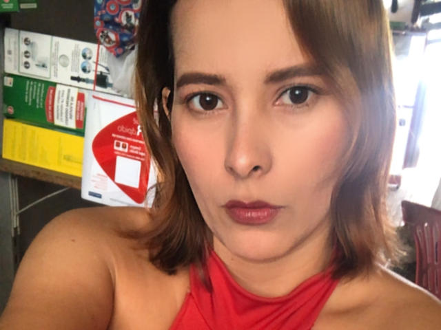 Sarita Velez mit braunen Haaren, einem roten Halbschultertop und rotem Lippenstift, die mit einem ernsten Gesichtsausdruck die Lippen zusammenpresst und dabei ihre nackten Schultern und dezentes Make-up zeigt..