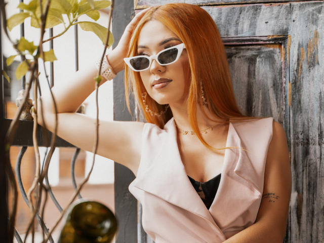 Maisy Warner, con lunghi capelli rossi, occhiali da sole bianchi, un gilet rosa, una maglietta nera, un tatuaggio sul braccio, una collana d'oro, un braccialetto e degli orecchini, appoggiata a una porta grigia e consumata..