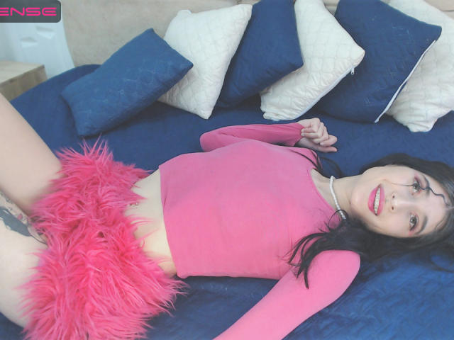 EvelinGonzales lies on a blue bed, wearing a pink crop top and fluffy pink skirt, exposing her tattooed pussy. She has black hair, green eyes, and a nose ring.