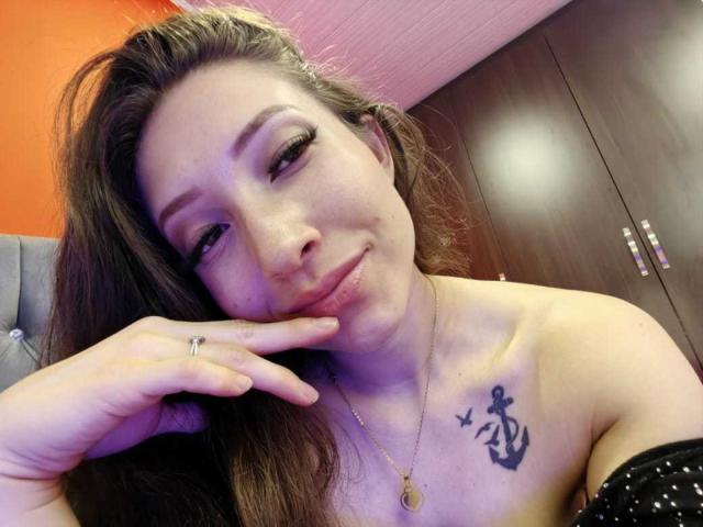 ElizabethMorrison has a tattoo of an anchor with a bird on her right tit. She's smiling with long brown hair, a ring on her finger, and a gold necklace.