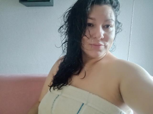 SakotoDika with wet black hair, looking at the camera, has small tits covered by a white towel, sitting on a pink couch, slightly smiling.