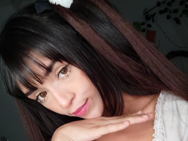 YourKatt has dark brown hair in pigtails with white hair ties, brown eyes, pink lipstick, and is smiling gently with her hand near her chin, wearing a white top.