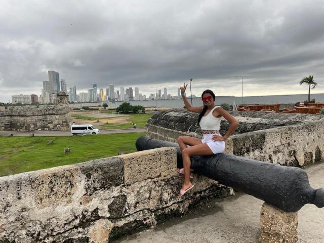 AzumiSmith with long black hair, red sunglasses, white top, and white shorts, sitting on a black cannon, making a peace sign, city skyline in background, cloudy sky.
