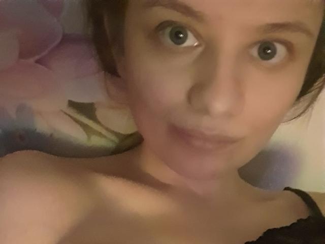 LinNatty hat große, blaue Augen, helle Haut und braunes Haar, sie lächelt mit leicht geöffneten Lippen und trägt schwarze Träger, wobei ihre nackten Schultern sichtbar sind. Ihr Gesicht ist im Bild zentriert..