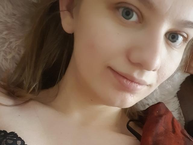 LinNatty hat blaue Augen, helle Haut und ein leichtes Lächeln. Sie trägt eine schwarze Spitzenbluse, und in der Nähe ihres Schulters befindet sich ein rotes Objekt. Ihr braunes Haar ist zu Zöpfen gebunden..
