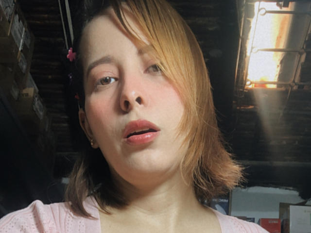 SaritaVelez has light brown, shoulder-length hair, pink lips, and fair skin. Her face is slightly blurred, with sunlight in the background. She's wearing a pink shirt.