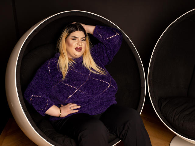 HornyBbw with blonde hair, purple sweater, black pants, and red lipstick, sitting in a round, black, upholstered chair, hand in hair, looking confident. Boobs are average size.