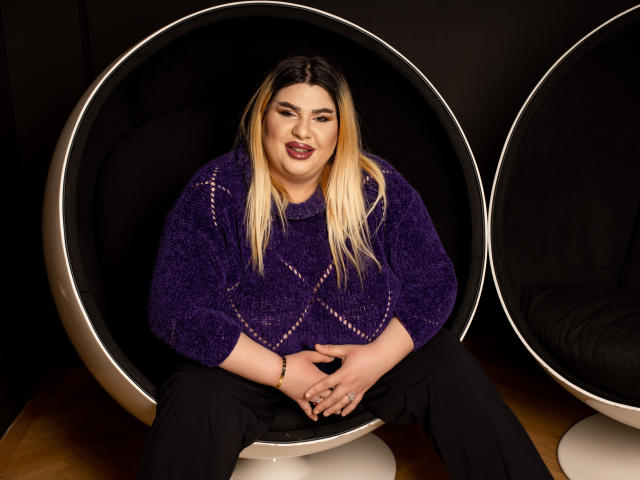 Une femme HornyBbw aux cheveux blonds, portant un pull violet et un rouge à lèvres foncé, est assise dans un fauteuil bulle noir et rond, les jambes écartées, les mains sur les genoux, en pantalon noir, sur un sol en bois, et elle sourit..