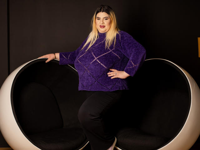 HornyBbw met blond haar, een paarse gebreide trui, zwarte broek, zittend in een ronde, zwarte, moderne stoel met haar hand op de rand van de stoel, kijkend naar de camera..
