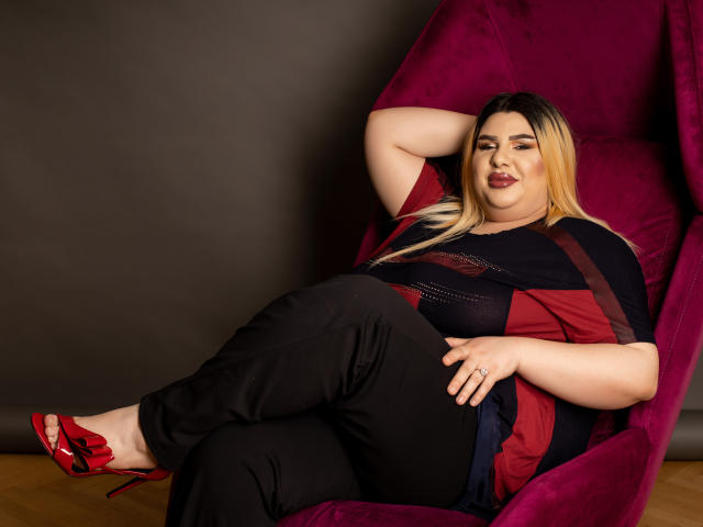 HornyBbw, une femme de grande taille avec des cheveux blonds et noirs, du rouge à lèvres rouge, une chemise noire et rouge, un pantalon noir, des talons rouges, assise confortablement dans un fauteuil violet, une main sur la tête, détendue, confiante..