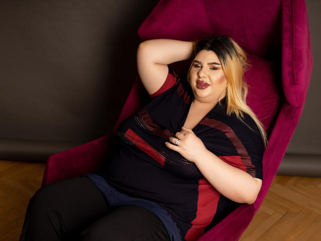 HornyBbw, een volslanke vrouw met blond haar en donkere aanzet, zit in een roze stoel, met haar hand op haar borst, en draagt een zwart en rode top. Ze kijkt er lichtelijk geïrriteerd uit..