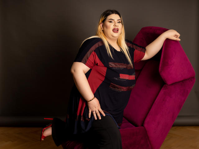 HornyBbw, curvy with large tits, sits on a magenta chair, wearing a dark, sheer top, black pants, and red heels, with blonde and black hair, and a sexy expression.