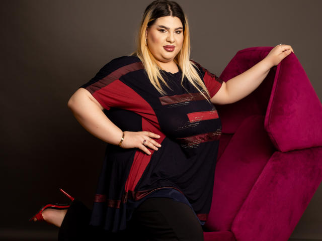 HornyBbw met blond haar, gekleed in een zwart en rood gestreept top, zwarte broek en rode hakken, zittend op een magenta stoel, hand op de heup, met een zelfverzekerde glimlach..