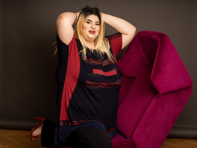 HornyBbw with blonde hair, red lipstick, and dark makeup, wearing a red and black dress, sitting on a pink velvet chair, arms raised, looking sexy, with red heels.
