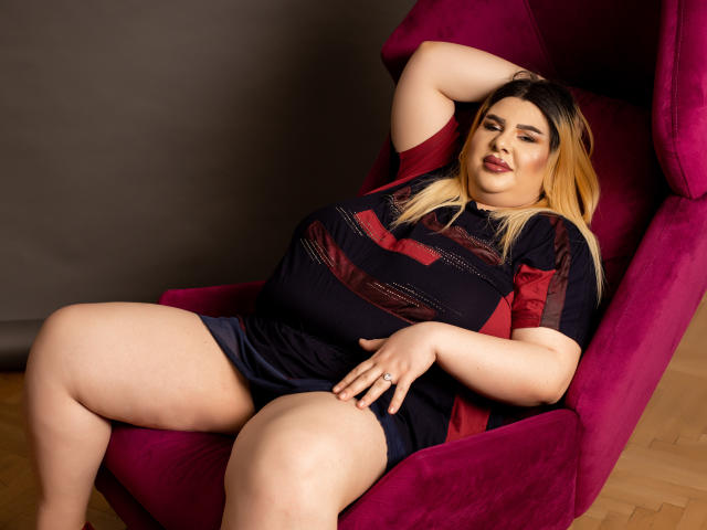 Une femme séduisante aux cheveux blonds, avec de grandes seins et des fesses rebondies, est assise dans un fauteuil magenta, vêtue d'une robe rayée noire et rouge, sa main sur son entrejambe, avec un sourire confiant..