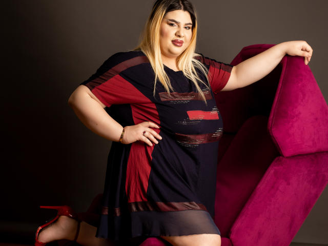 HornyBbw, femme corpulente aux cheveux blonds, rouge à lèvres rouge, robe noire et rouge, talons hauts rouges, appuyée sur une chaise violette, confiante, sexy, pulpeuse, voluptueuse, debout dans un studio..