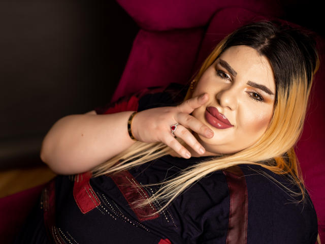 HornyBbw, une femme corpulente avec des cheveux blonds et noirs, fait la moue en plaçant sa main près de ses lèvres roses, vêtue d'une robe noire et rouge, assise sur une chaise rose..