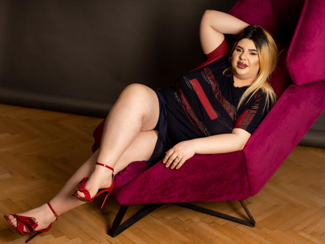 Une femme séduisante aux cheveux blonds, au rouge à lèvres rouge, portant une robe noire transparente, des talons rouges et assise sur un fauteuil en velours rouge, montrant ses seins et ses fesses, posant avec une main derrière la tête..