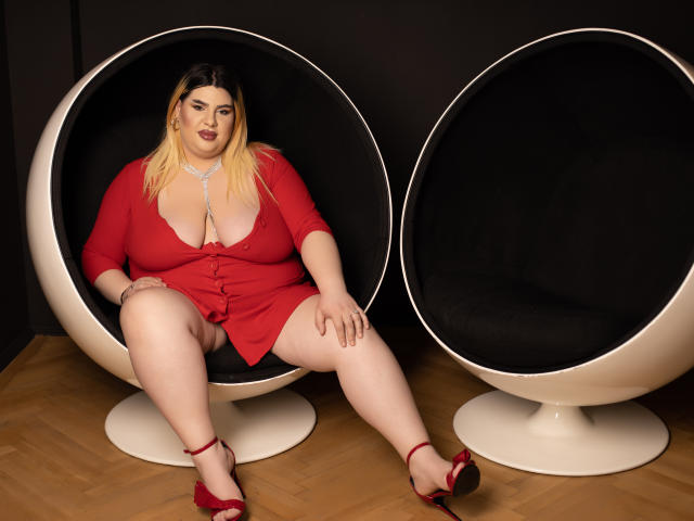 HornyBbw with big tits, pale skin, blonde hair, red dress, and red heels sits in a black egg chair, legs spread, showing her pussy. Another empty egg chair on right.