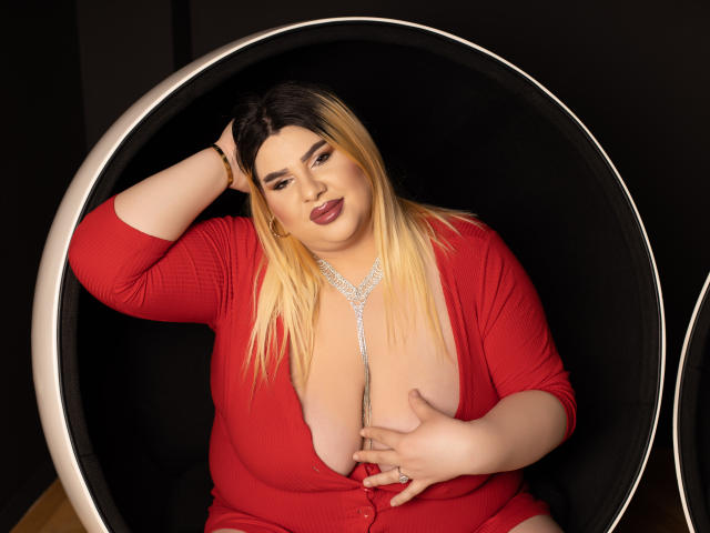 Une femme séduisante avec de grandes seins, portant un cardigan rouge, assise dans un fauteuil noir sphérique, une main sur la poitrine, cheveux blonds, sourcils foncés, rouge à lèvres rouge et un collier en argent..