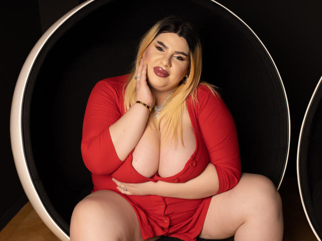 HornyBbw met blond en zwart haar, grote borsten en een rode jurk, zittend in een zwarte eierstoel, hand op de wang, benen gespreid, kijkend naar de camera met een zelfverzekerde glimlach..