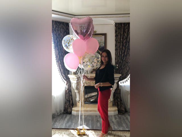 MaryLoveX with pink, white, and heart-shaped balloons, wearing a black top and red pants, standing in a room with a white fireplace and dark curtains.