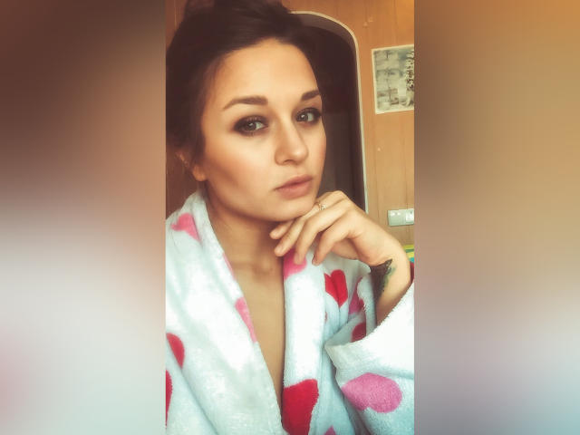 MaryLoveX, portant un peignoir rose et blanc duveteux, avec ses cheveux bruns attachés en un chignon désordonné, ses yeux verts, un maquillage discret et une expression pensive, appuyant son menton sur sa main..