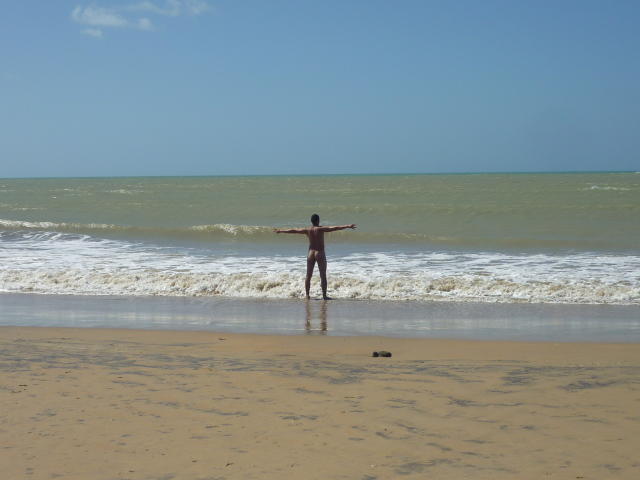 AlexMllo staat op een strand met gespreide armen, naar de oceaan gericht, en draagt rode zwemkleding. Hij heeft een donkere huidskleur. Zijn rug is naar de camera gericht..