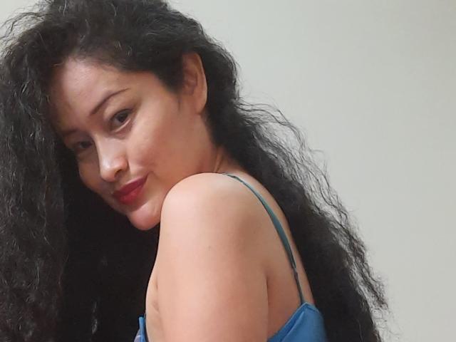 Xuuley, con cabello negro rizado, lápiz labial rojo y un vestido azul con tirantes, sonríe levemente, mostrando un hombro descubierto, y mira por encima del hombro. Sus senos son visibles, mostrando un aspecto lindo y sexy..