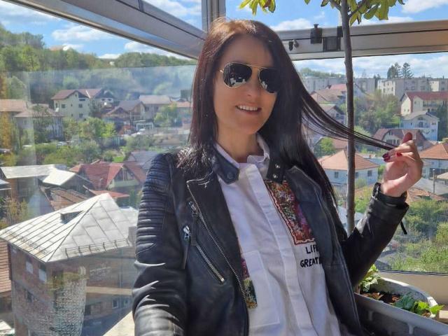 JoliDanielle with long black hair, wearing sunglasses, a black leather jacket, white shirt, and colorful necktie, holding a cigarette, standing by a window with a city view.