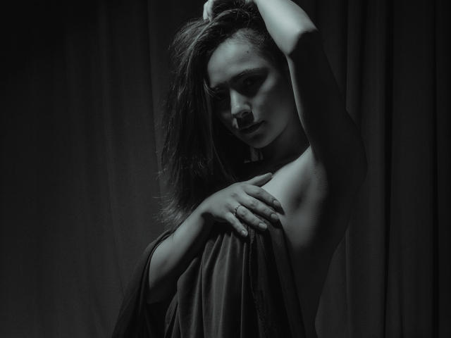 ValeryHarllow with dark hair, holding a sheer cloth over her tits, arms raised, shadowy green-black lighting, seductive expression, hand on chest, soft texture, moody atmosphere.