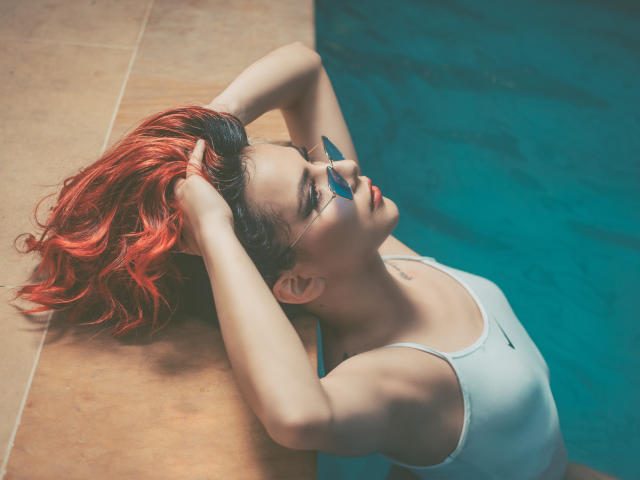 ValeryHarllow with red hair, hands in her hair, wearing glasses and a white sports bra, leaning against a pool edge. Her blue eyes are closed, showing a relaxed face.