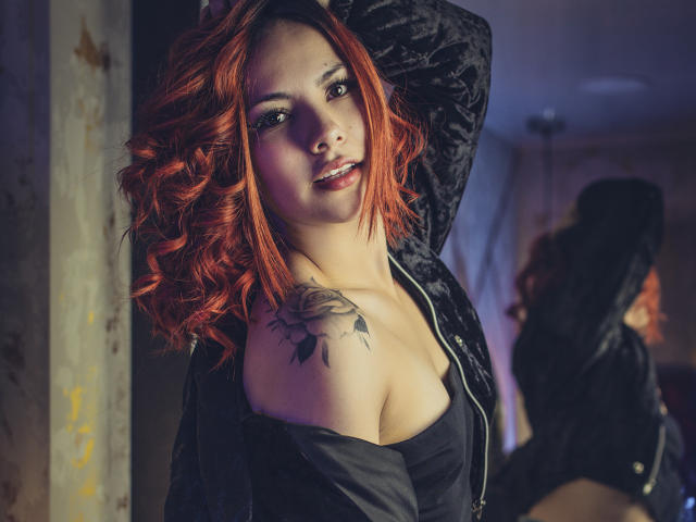 ValeryHarllow with red curly hair, black off-shoulder top, and rose tattoo on right shoulder, arm raised, looking at camera, soft light, subtle smile, mirror reflection in background.