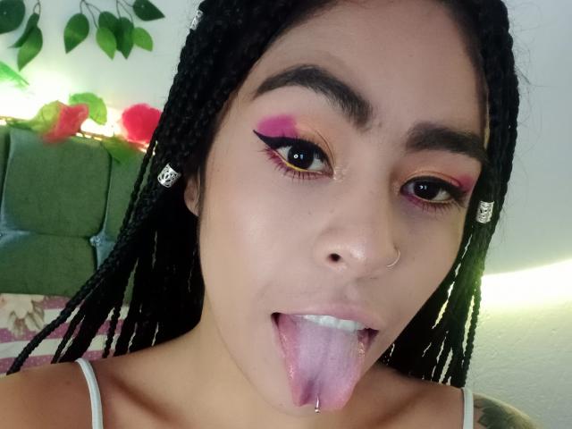 StellaBeckker's face with braided hair, pink and orange eyeshadow, black eyeliner, nose ring, tongue out, drooling, and a pierced tongue, wearing a white top.