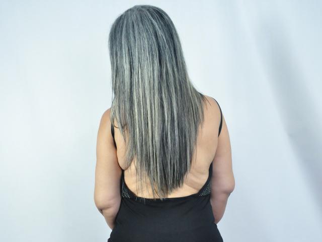 GabrielleMature, con la schiena rivolta alla telecamera, capelli grigi e argentati, vestito nero a spalline sottili, braccia leggermente dietro, schiena scoperta e spalline nere visibili..
