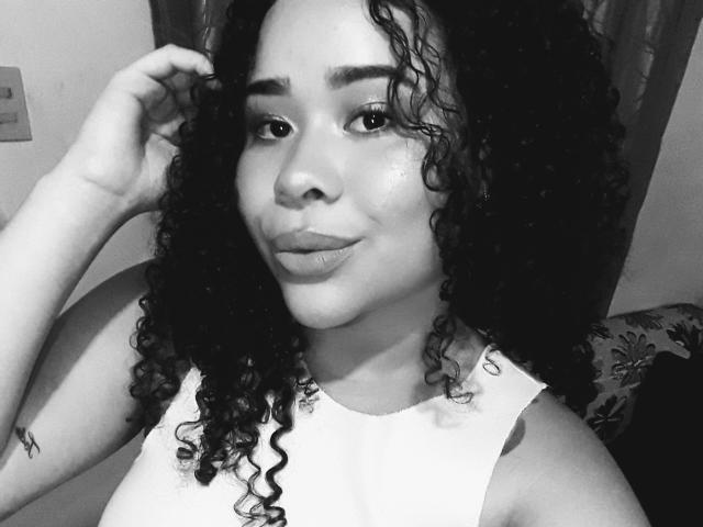 Foto em preto e branco de EveNailah com cabelo cacheado, lábios carnudos, pele clara, vestindo uma blusa branca sem mangas, com a mão no cabelo, uma pequena tatuagem no braço esquerdo e um sorriso discreto.