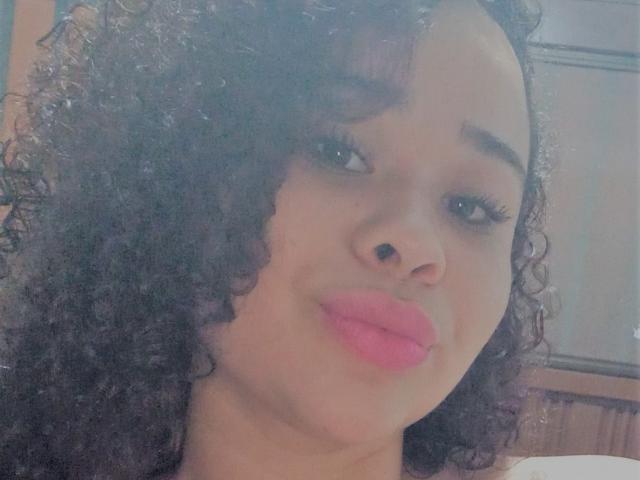 EveNailah, met krullend zwart haar, roze lippenstift en bruine huid, kijkt naar de camera met een lichte glimlach, waarbij haar neus en volle lippen zichtbaar zijn. Haar wimpers zijn lang..