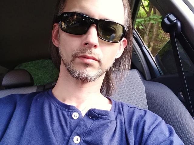 Latinx69 with a goatee, wearing black sunglasses, and a blue button-up shirt, sitting in a car with gray patterned seat. Long brown hair, serious expression, green car seat.