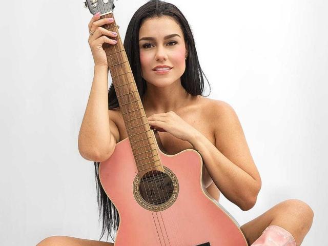 SlinkyAngeel mit langen, schwarzen Haaren, die eine pinke Akustikgitarre hält, oben ohne sitzend, in die Kamera schauend, lächelnd, mit den Fingern auf den Saiten, pinke Socken, leicht gebräunt, weiches Licht, entspannte Pose..