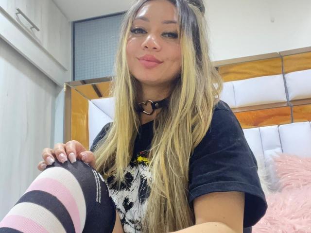 SophieLombardo with long blonde hair, black choker, black shirt, pink and black striped thigh-highs, sitting on bed, pink fluffy pillow, smiling, pink lipstick, and pink nail polish.