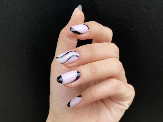 Pinglis Hand mit einem blassrosa und schwarzen, wellenförmigen Nagellackdesign, schwarzen Spitzen und spitzen Nägeln, vor einem schwarzen Hintergrund..