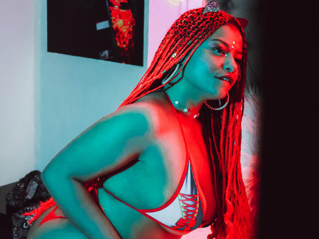 NashaJonny with red braided hair, large boobs in a white and red lace-up bikini, green and red light on her, large hoop earrings, looking to the side with a smile.