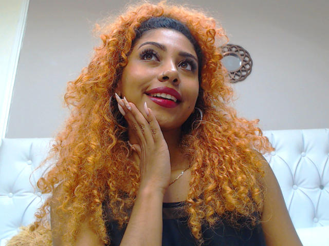 LilithWalker with orange curly hair, brown eyes, red lipstick, and black sleeveless top, smiling, hand on cheek, white tufted couch in background, ring on right hand.